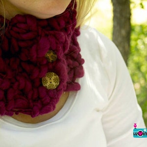 May include: A close-up of a woman wearing a deep burgundy knitted cowl neck scarf with decorative wooden buttons. The scarf is made of a soft, chunky yarn.