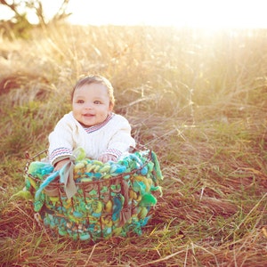 May include: A baby in a white sweater sits in a woven basket with green, blue, and brown accents. The baby is smiling and looking to the right. The basket is sitting in a field of tall grass.