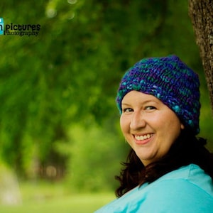 May include: A woman with long brown hair is wearing a blue and purple knitted hat. She is smiling and looking at the camera. The hat is a chunky knit with a textured pattern.