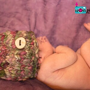 May include: A newborn baby wearing a knitted hat with a wooden button on a purple background. The baby is sleeping with their arms tucked in and their legs crossed.