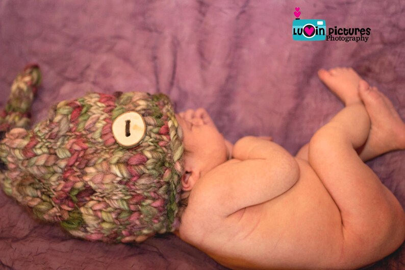 May include: A newborn baby wearing a knitted hat with a wooden button on a purple background. The baby is sleeping with their arms tucked in and their legs crossed.