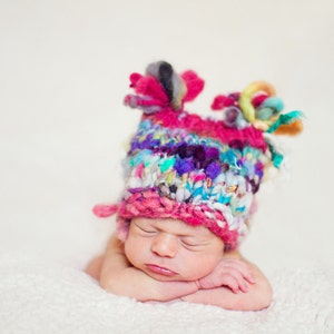 May include: A newborn baby wearing a colorful knitted hat with pink, blue, white, and purple yarn. The baby is sleeping with their arms crossed under their chin.