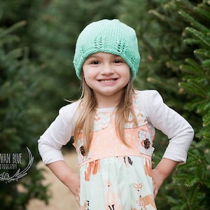 May include: A young girl wearing a light green knitted hat with a white long-sleeved shirt and a floral print dress. She is standing in front of a green evergreen tree.