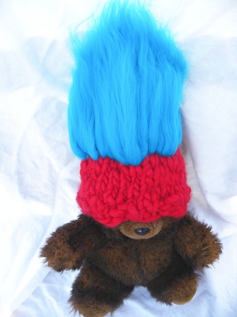 May include: A brown teddy bear wearing a red knitted hat and a bright blue wig. The hat covers the bear's eyes.