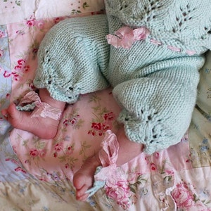 Puede incluir: Un bebé con un atuendo de punto azul claro con detalles de cinta rosa. El bebé está acostado sobre una colcha con estampado floral.