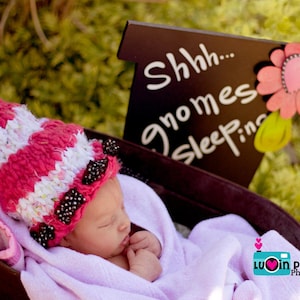 Peut inclure: Un bébé portant un bonnet tricoté rayé rose et blanc avec des perles noires et une couverture rose, dormant dans un berceau en bois. Le berceau porte une pancarte qui dit "Shhh... gnomes sleepin".