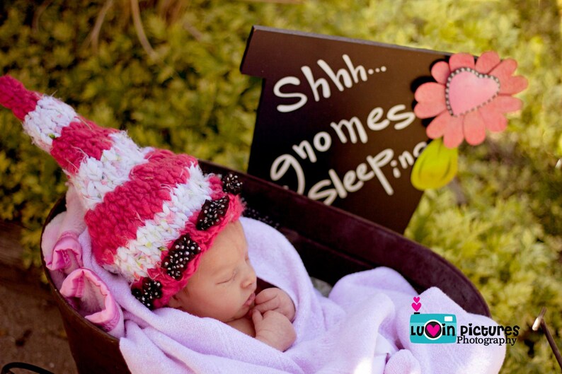 May include: A pink and white striped knitted hat with black beads is on a baby's head. The baby is lying in a white blanket in a wooden crib. A sign above the baby reads "Shhh... gnomes sleepin".