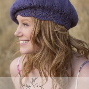 以下が含まれることがあります： 長く波打つ明るい茶色の髪をした女性が、レースの縁取りがついた紫色のニットのベレー帽をかぶっています。彼女は微笑んで横を向いています。彼女は花柄の白いタンクトップを着ています。