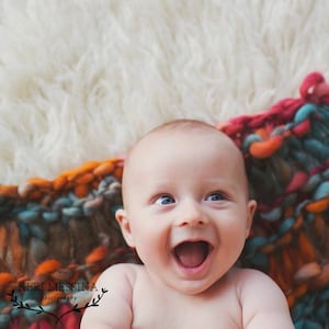 May include: A baby with bright blue eyes is lying on a colorful knitted blanket. The baby is smiling and has a happy expression on their face.