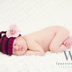 May include: A sleeping newborn baby wearing a purple and brown striped knitted hat with a pink flower on top. The baby is lying on a white fluffy blanket.