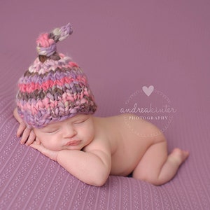 May include: A newborn baby wearing a pink, purple, and brown knitted hat with a knot on top, sleeping on a purple textured blanket. The baby is lying on their side with their head resting on their arm.