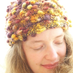 May include: A woman wearing a colorful knitted hat with a textured, bumpy design. The hat is made with a variety of shades of brown, yellow, and purple yarn.