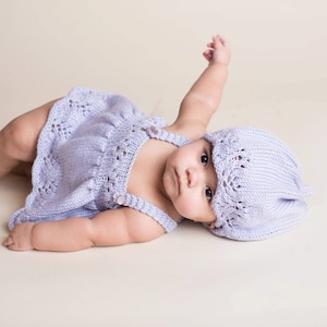 Op de afbeelding: Een baby die een lavendelkleurige gebreide jurk en bijpassende muts draagt. De baby ligt op zijn rug met één arm omhoog.