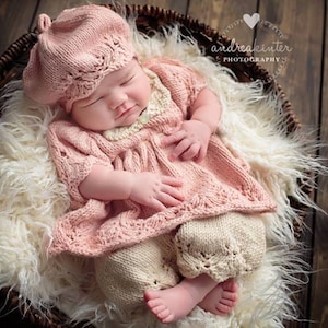 Op de afbeelding: Een baby die een roze gebreide jurk en bijpassende baret draagt, slaapt in een rieten mand bekleed met wit bont.