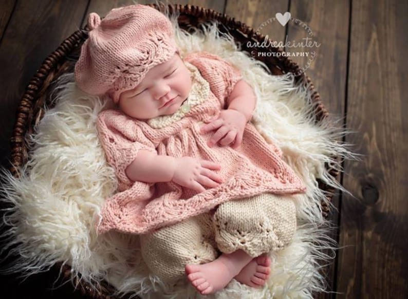 Op de afbeelding: Een baby die een roze gebreide jurk en bijpassende baret draagt, slaapt in een rieten mand bekleed met wit bont.