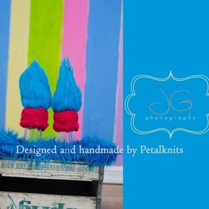 May include: Two blue and pink knitted troll doll hats on a wooden crate with a blue fuzzy surface. The background is a colorful striped wall. Designed and handmade by Petalknits.