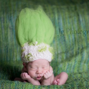 May include: A newborn baby wearing a green and white knitted hat with a green fluffy top. The baby is lying on a green and blue textured surface.