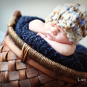 May include: A newborn baby wearing a brown, white, and blue knitted hat is sleeping in a woven basket lined with a blue blanket.