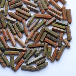 Könnte beinhalten: Eine Sammlung von Unakit-Kristallstäben, jeweils sechseckig und mit spitzer Spitze. Die Stäbe zeigen eine Mischung aus Farben, darunter Grüntöne, Orange und Braun. Die Kristalle sind in einem kreisförmigen Muster vor einem weißen Hintergrund angeordnet.