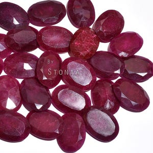 May include: A collection of oval-shaped, faceted ruby gemstones in varying shades of red. The gemstones are arranged closely together, with the text "BY STONAA" visible on one of the gems. The background is white.