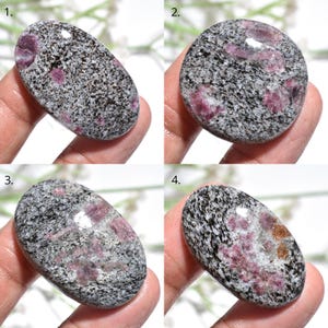May include: Four close-up images of polished gemstones. The stones have a speckled grey and black base colour with patches of pink and reddish-brown. The shapes vary from oval to round. The gemstones are held in a hand.