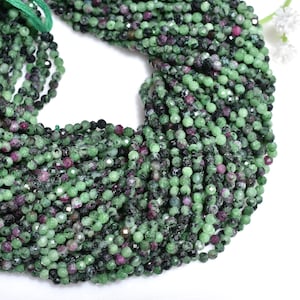May include: Close-up of a strand of faceted beads. The beads are a mix of green, black, and red. The beads are strung together with a green string. The background is white.