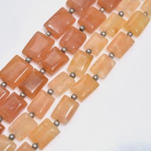 May include: Close-up of a necklace featuring rectangular, peach-colored gemstones strung together with small, silver-colored beads. The gemstones have a polished, translucent appearance, and the necklace is set against a white background.