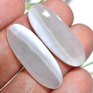 May include: Two oval-shaped, smooth, polished cabochons featuring a translucent, milky white appearance with a distinct, opaque, light gray horizontal band running across the center of each stone, showcasing a natural, layered mineral texture.