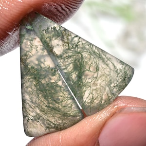 May include: A translucent, triangular moss agate gemstone with intricate green, fern-like inclusions. The stone is held between fingers, showcasing its natural patterns and clarity. The gem's polished surface reflects light, highlighting its unique internal structures.