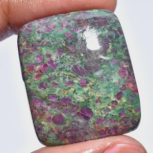 Pode incluir: Uma pedra preciosa retangular com uma mistura de cores. A pedra tem uma base verde com numerosas inclusões rosa escuro e roxo. A superfície é polida, refletindo a luz. A aparência geral é natural e texturizada.