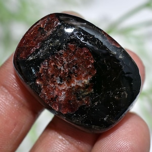 May include: A polished gemstone with a mix of black and reddish-brown colors. The stone has a smooth, rounded shape and a glossy surface. The reddish-brown areas have a crystalline appearance, while the black areas are more opaque.