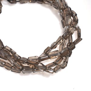 May include: Close-up of a necklace composed of rectangular, smoky quartz beads. The beads are strung together, forming multiple strands. The beads' colour is a translucent, brownish-grey, with a polished finish. The necklace is shown against a white background.