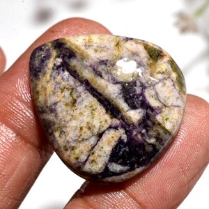 May include: A polished, irregularly shaped gemstone with a mix of colors. The stone features a base of creamy white and tan, with streaks of deep purple and patches of green. The surface has a glossy sheen.