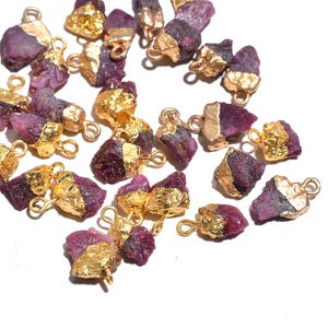 Könnte beinhalten: Eine Sammlung roher Rubinanhänger mit vergoldeten Kanten und Schlaufen zur Schmuckherstellung. Die Anhänger haben eine tiefrote bis violette Farbe und eine raue, natürliche Textur. Die Vergoldung verleiht den rohen Edelsteinen einen Hauch von Eleganz.
