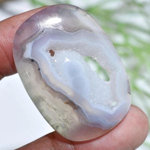 Può includere: Una pietra di agata ovale lucidata con un centro di geode. La pietra presenta strati di tonalità bianche, grigie e lavanda. Il centro della geode è una cavità riempita di cristalli bianchi scintillanti. La pietra è tenuta tra le dita.