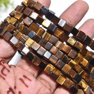 May include: Close-up of strands of square tiger eye beads. The beads are a mix of brown, gold, and black, with a polished, reflective surface. The beads are held in a hand, showcasing their natural variations.