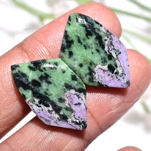 Pode incluir: Duas pedras preciosas polidas com um padrão único. As pedras apresentam uma cor de base verde claro com inclusões pretas e roxas. As pedras preciosas são seguradas em uma mão, mostrando sua beleza natural e detalhes intrincados.