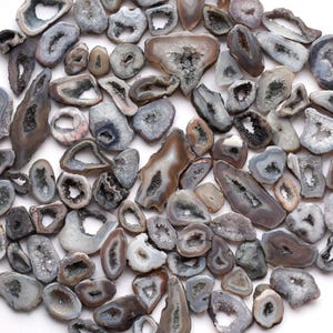 May include: A selection of polished agate geodes in various sizes and shapes, featuring grey, brown, and white hues. The geodes display hollow centres with visible crystal formations. The arrangement is scattered on a white surface, highlighting the natural beauty of the stones.