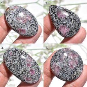 Puede incluir: Cuatro piedras preciosas pulidas con una matriz moteada gris y negra. Cada piedra presenta parches de cristales de color rojo rosado. Las formas incluyen ovalada, de lágrima y redonda. Las piedras preciosas se sostienen en una mano.