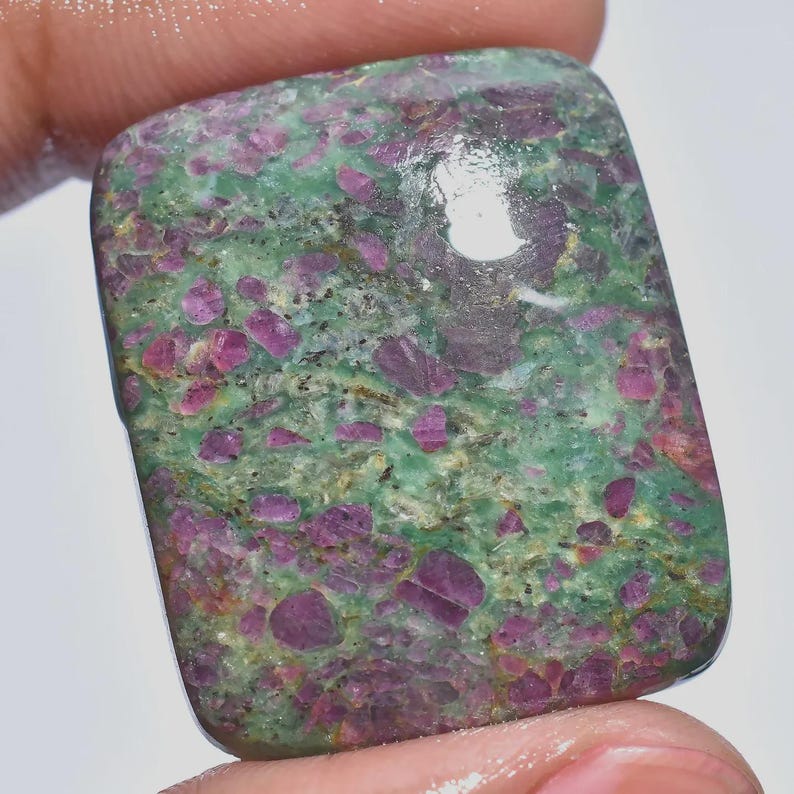 May include: A rectangular gemstone with a polished surface. The stone features a mottled pattern of deep pink and purple inclusions set in a matrix of green. The gemstone is held between fingers.