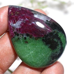 Könnte beinhalten: Ein polierter, tropfenförmiger Edelstein mit einer auffälligen Farbkombination. Der Stein zeigt einen satten, tiefroten Farbton, der in ein leuchtendes Grün übergeht, mit Bereichen in Schwarz und Weiß. Der Edelstein ist glatt und reflektierend und zeigt natürliche Muster.
