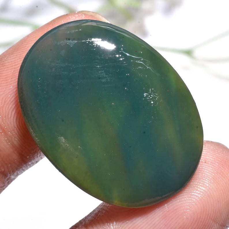 May include: Oval-shaped gemstone with a polished surface. The stone features a blend of green and blue hues, with subtle color variations. The gemstone is held between fingers, showcasing its size and shape. Ideal for jewelry making.