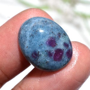 Op de afbeelding: Een gepolijste, ovaalvormige edelsteen met een blauwgrijze basiskleur en levendige paarse insluitsels. De steen heeft een glad, glanzend oppervlak, met kleine donkere spikkels. De edelsteen wordt tussen twee vingers gehouden.