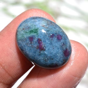 May include: A smooth, oval gemstone with a blue-green hue. The stone has dark red and green inclusions, creating a unique pattern. The gem is held between fingers, showcasing its smooth, rounded form.
