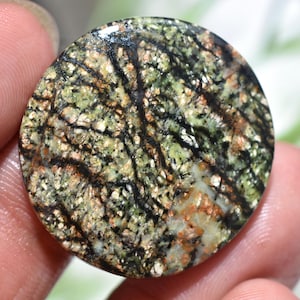 May include: A round, polished gemstone with a unique pattern. The stone features a mix of green, brown, and black colors, with a network of dark lines across the surface. The overall appearance is reminiscent of a natural landscape.