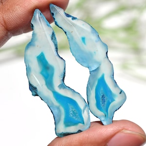 May include: Two polished agate slices with a natural, irregular shape. The stones feature a striking pattern of white, light blue, and deep turquoise, with a crystalline center. The edges are a darker blue, creating a beautiful contrast.