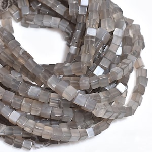 Puede incluir: Primer plano de cuentas grises en forma de cubo. Las cuentas son translúcidas y tienen un aspecto ligeramente pulido. Están ensartadas, formando un hilo flexible. Estas cuentas probablemente se utilizan para la fabricación de joyas u otros proyectos de artesanía.