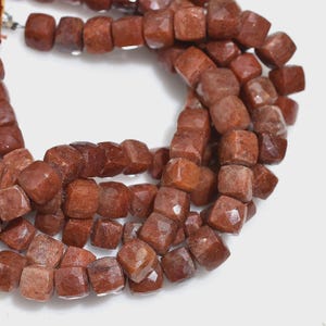 May include: A strand of faceted, cube-shaped sunstone beads in a deep reddish-brown hue. Each bead features a polished, multi-faceted surface that reflects light, arranged in a continuous loop on a thin, flexible string for jewelry making.