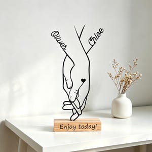 Puede incluir: Arte mural de metal negro que representa dos manos entrelazadas, con los nombres "Oliver" y "Chloe" escritos encima. Un pequeño corazón está entre las manos. El arte está montado sobre una base de madera que dice "Enjoy today!". Un pequeño jarrón blanco con flores secas está a la derecha.