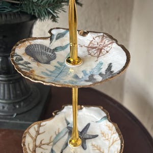 May include: A three-tiered serving tray with a gold-colored handle and support. The trays are shell-shaped, decorated with a nautical theme, including seashells, starfish, and coral. The edges are trimmed with gold.